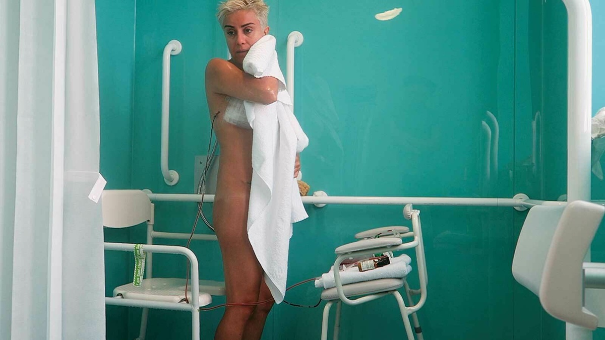 Mania standing naked post-surgery, holding a towel as she dries her face.