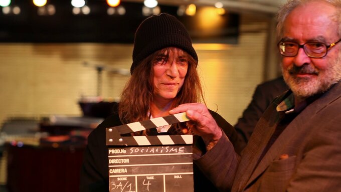 Patti Smith and Jean Luc Godard smiling at the camera, Godard is holding a film clapper with the word SOCIALISM on it.