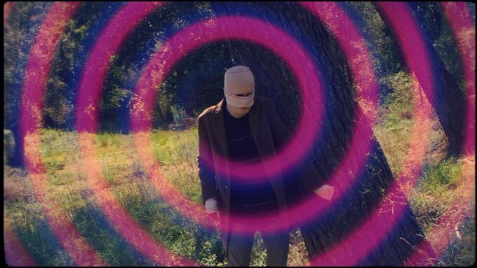 A man with a bandaged face walks in a forest. Around it we see a pink spiral that takes us to psychedelia.