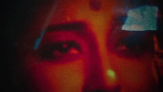 A woman's face fills the image in a red and orange glow, she wears a bindi and looks out with a yearning expression.