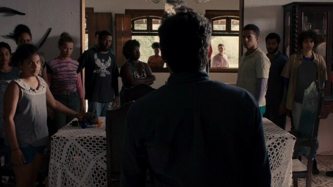 A man inside a rural mansion in Brazil is observed by several characters who look at him intimidatingly and intently.