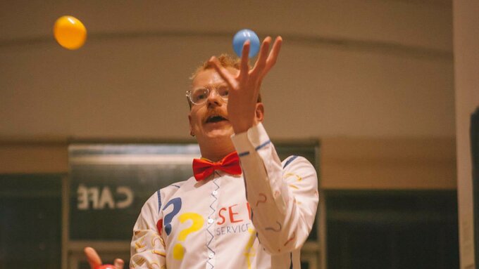 A white person with a white shirt juggling colourful balls performs in a cafe as a crowd watches and smiles.