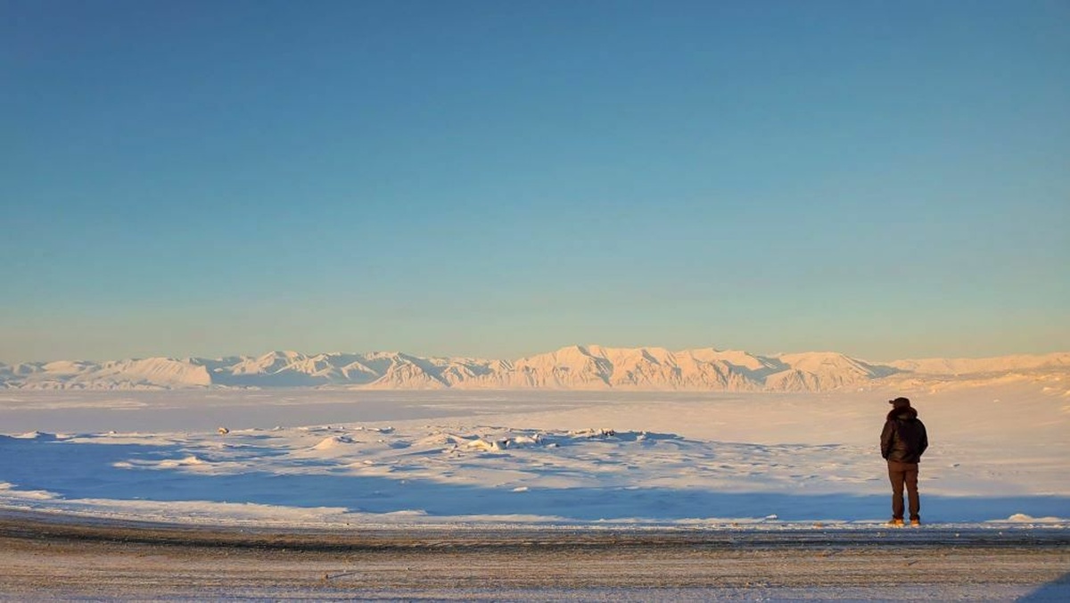 A snow-covered mountainous landscape against a blue sky. A person in a black puffer jacket looks towards the mountains.