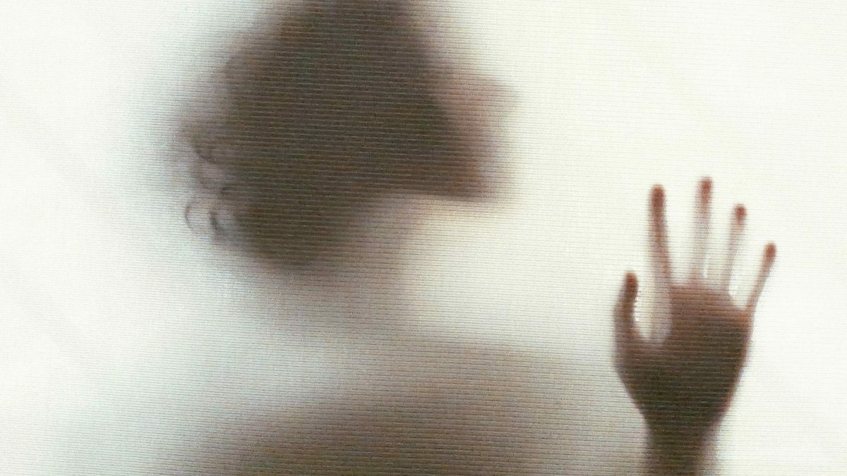 A blurred silhouette of a person seen through an opaque glass wall, their head and hand visible as they touch the pane.