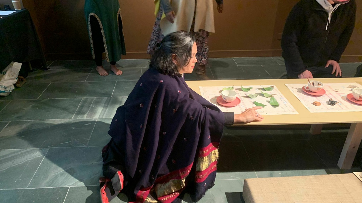 The artist, Annalee Davis sit's at a low table wearing a long purple and gold shawl, she points at various leaves.