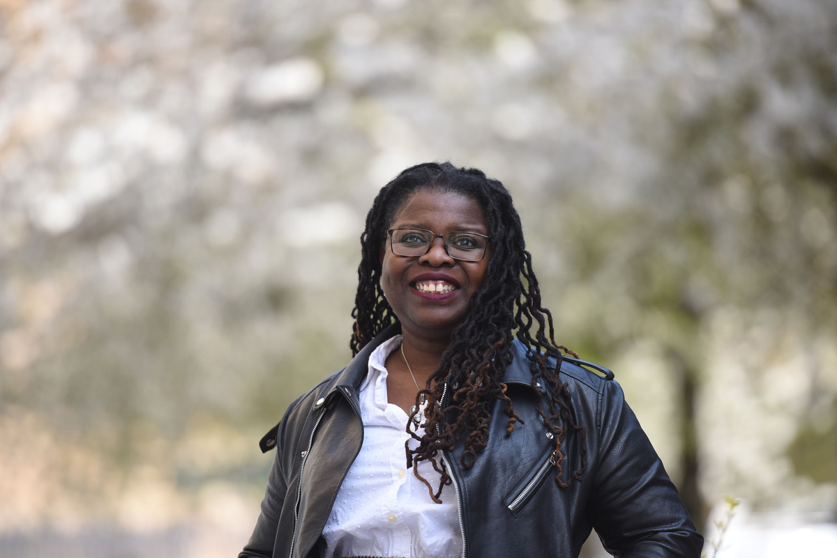 A smiling woman in the centre of the frame, she's wearing a black leather jacket, stylish glasses, and her hair in locks