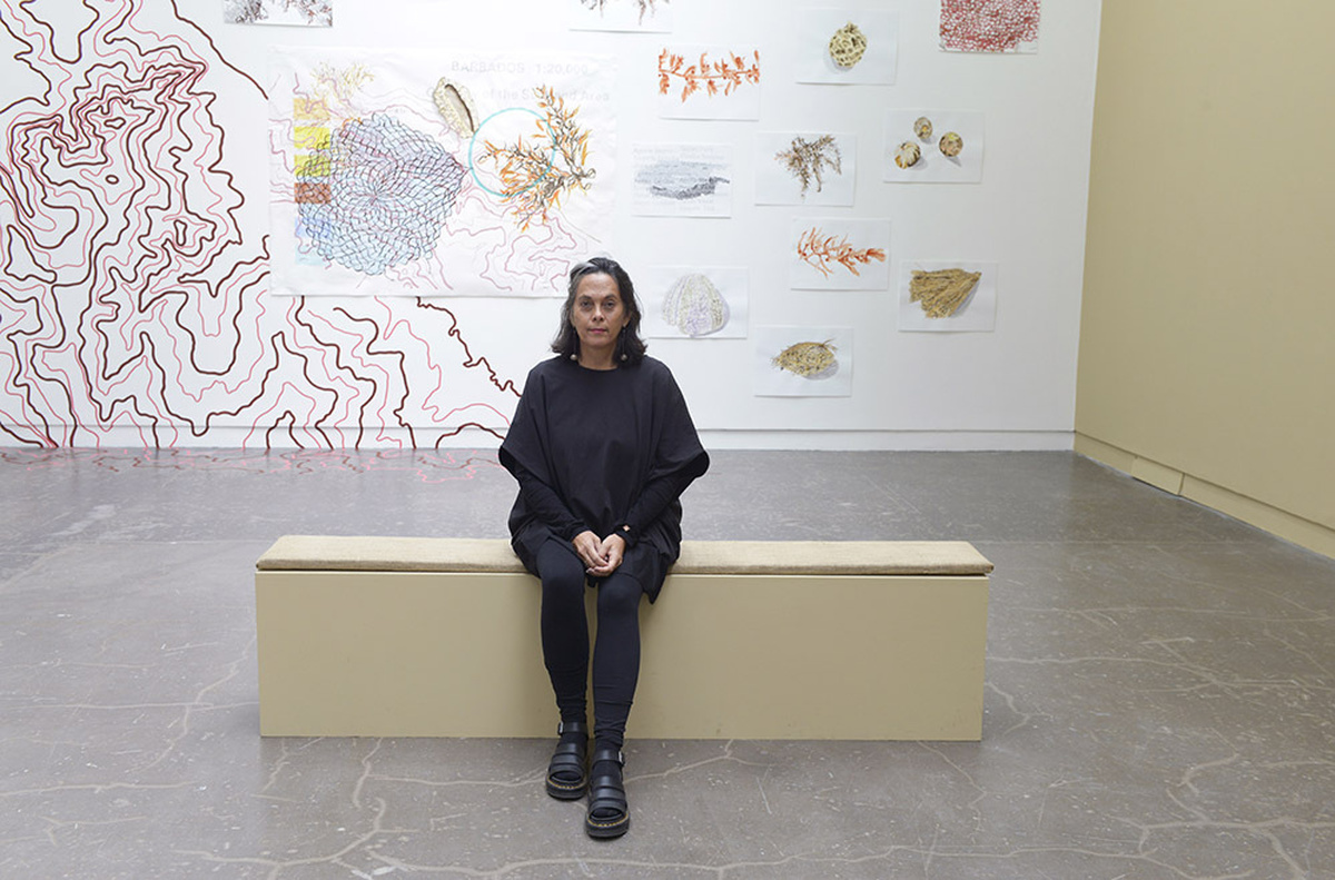 A woman sits on a bench hands clasped in a gallery space. She in all black, her hair is parted and has dangling earrings