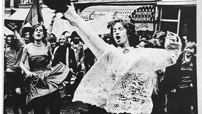 ﻿A black and white newspaper clipping of two gender nonconforming people marching joyfully.