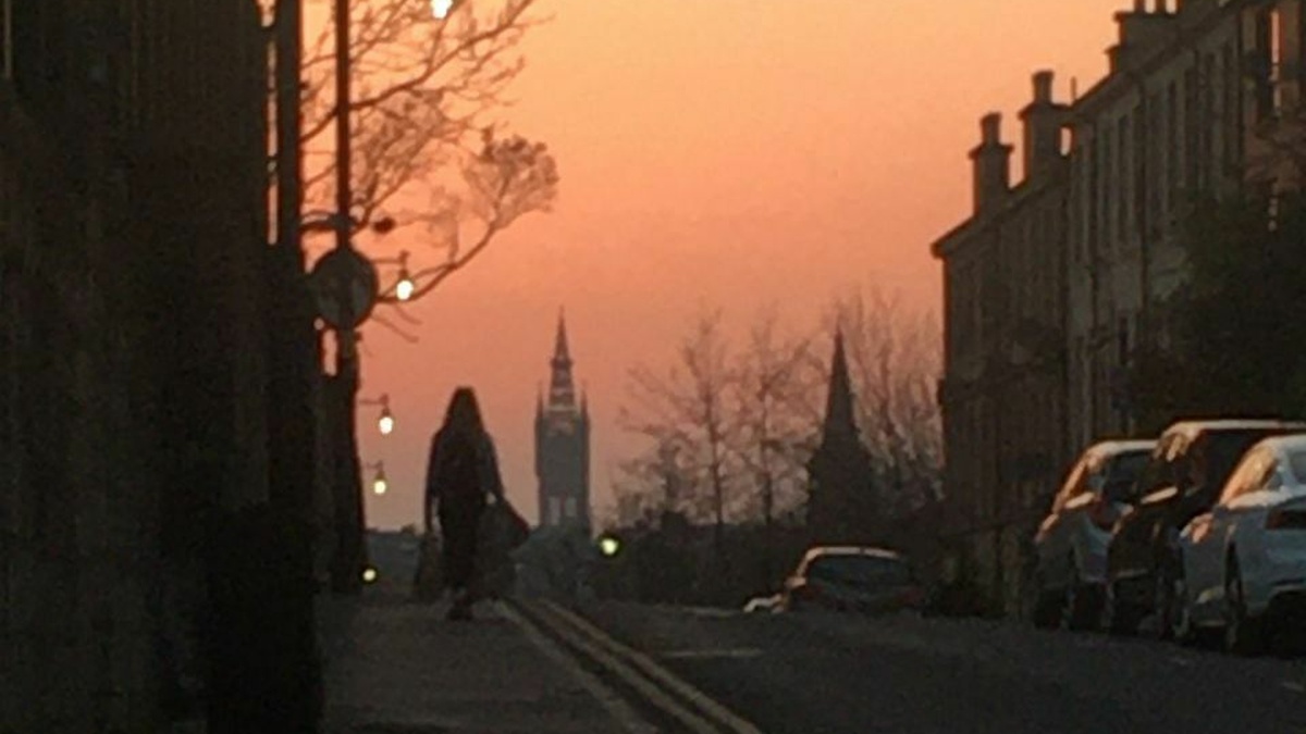 A silhouetted figure walking down a city street at dusk, the Glasgow University spire is visible in the distance.