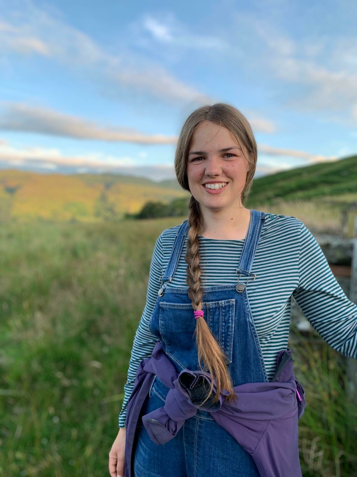Young storyteller, Ailsa Dixon, smiles at the camera against a background of blue skies and mountains.