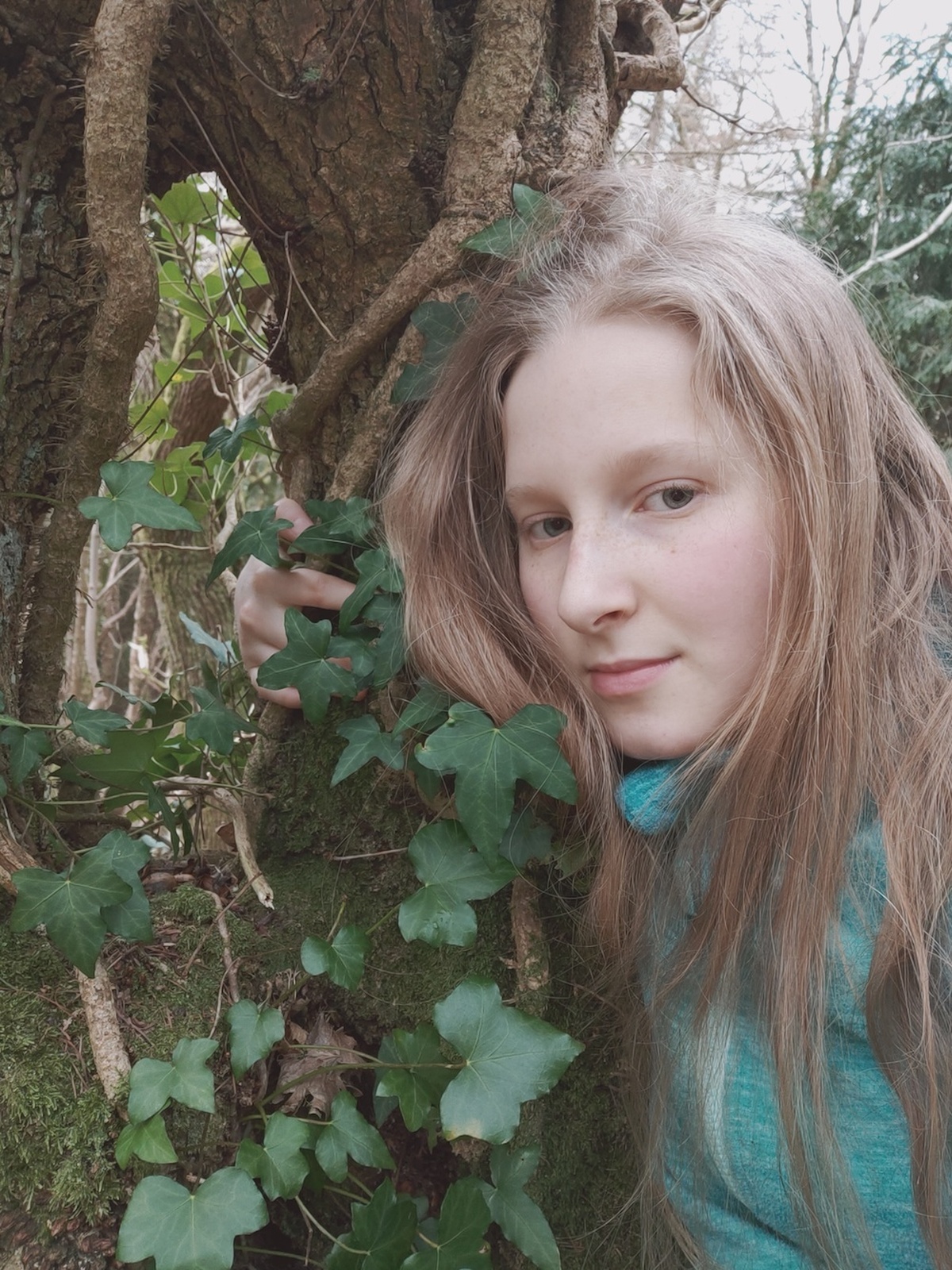 Young storyteller Ffion Phillips, looks towards us, leaning against and holding on to a tree trunk covered in ivy
