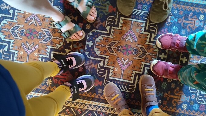 View from above of the legs and feet, with a range of colourful shoes of five people standing on a geometric carpet.