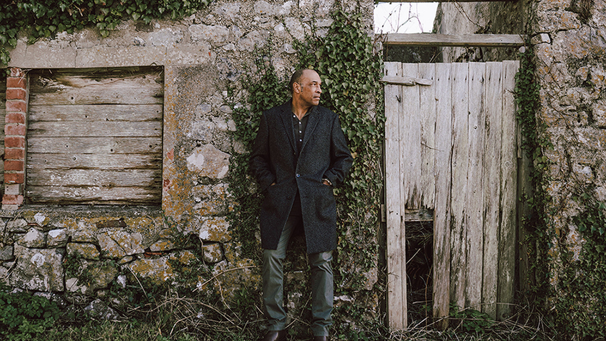 A smartly dressed man stands in front of dilapidated country house.