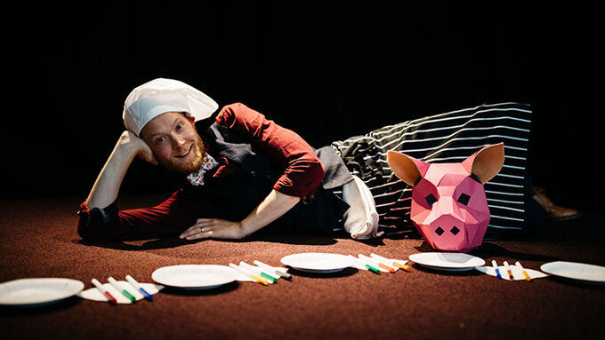 Dan Serridge wearing a white chef's hat, is lying on the floor with craft materials and a pink pig mask in front of him.