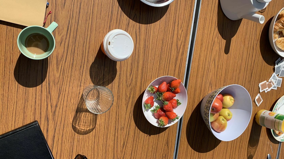 A brown trestle table from above. On the table a bowl of strawberries, coffe cups, apples, a teapot and a notebook.