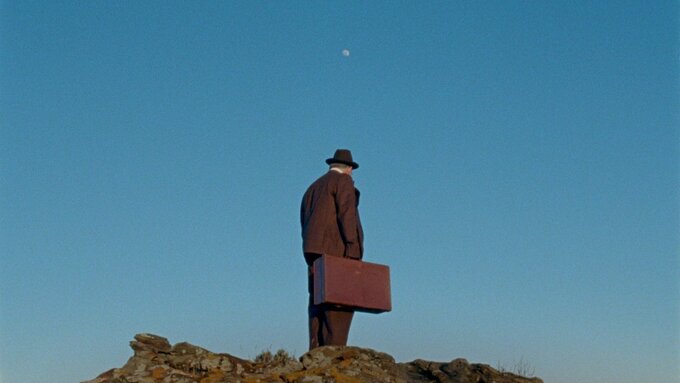 A man in a suit and trilby hat carrying a suitcase stands on a rocky hill. The full moon is seen high above him.