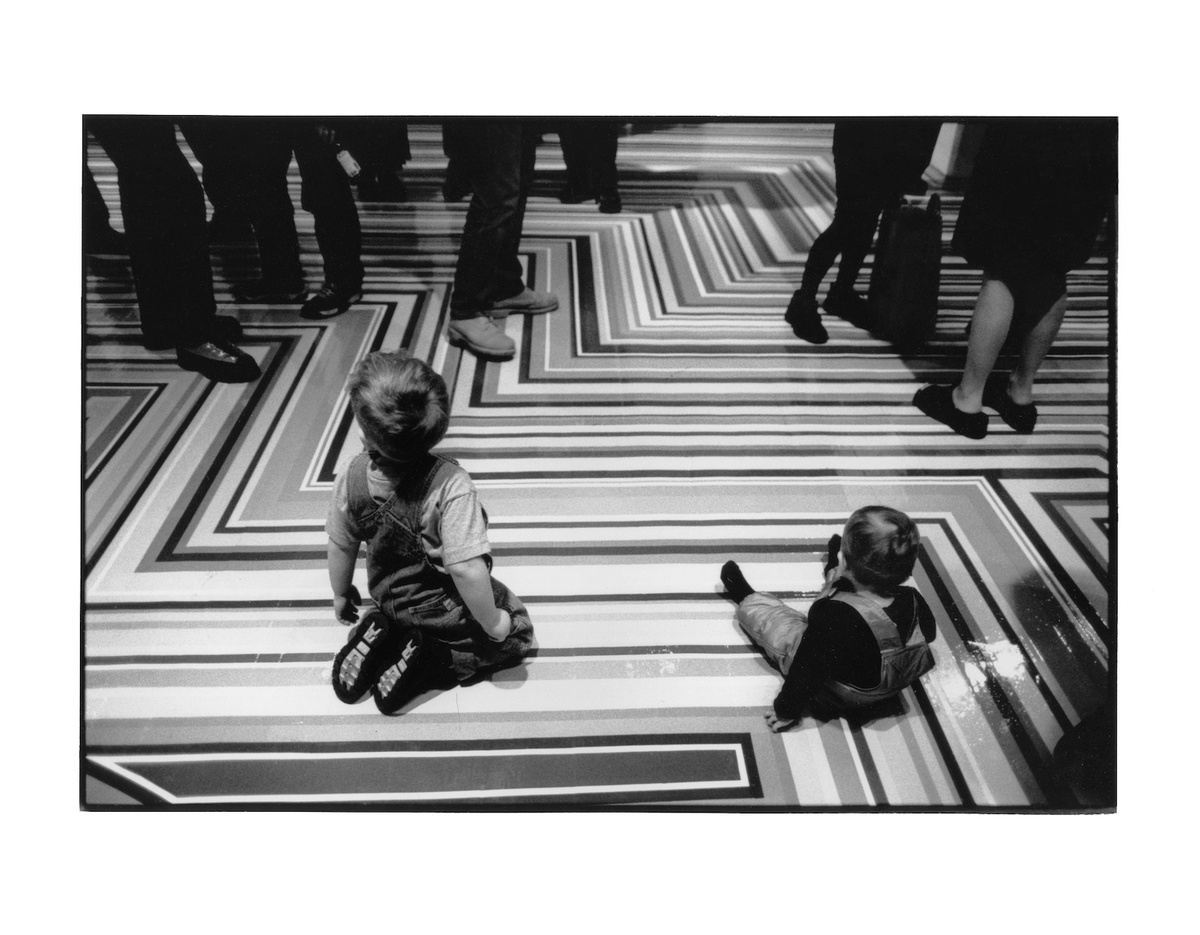 Two children sit on a Jim Lambie striped floor in the foreground with legs of gallery goers in the background.