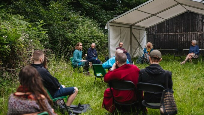 A group of people sat on camping chairs in a field outside with a gazebo and trees in the background