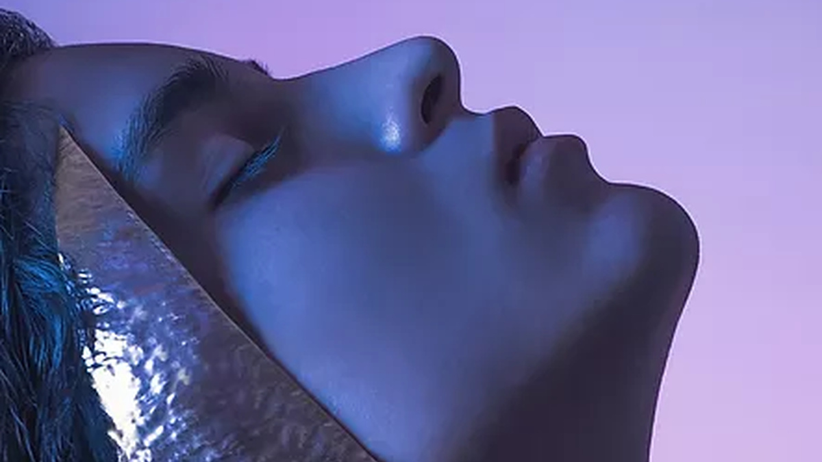 A head lit in profile looking serene, lit in purple and blue. With a gold sci-fi ornamentation beside their face.