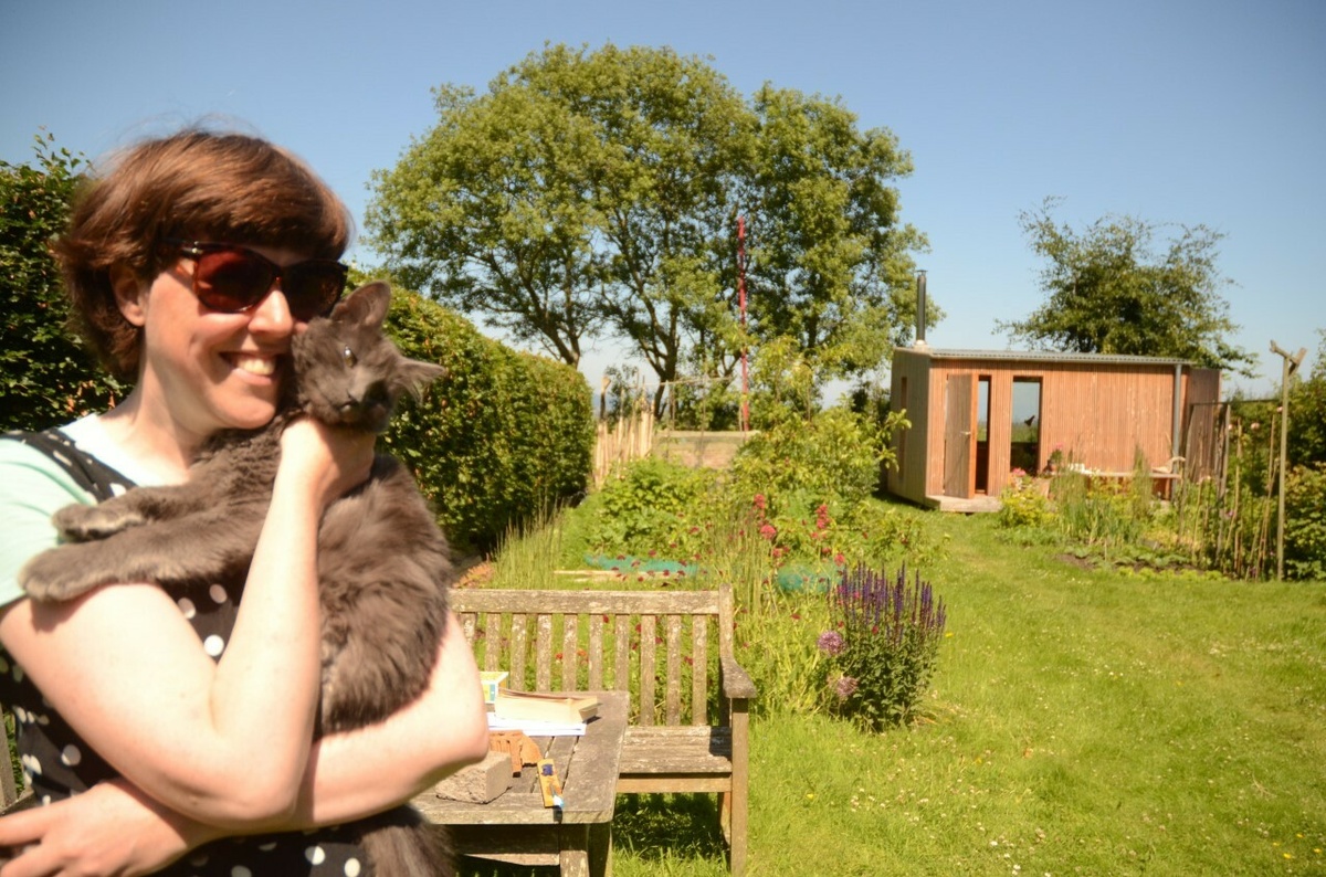 Kate Temple wearing sunglasses poses with Orlando the cat in a sunny, lush garden