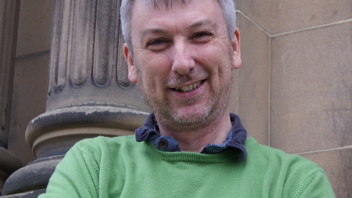 A photo of Tom Murray smiling with his arms folded, in front of a sandstone building, wearing a green jumper.