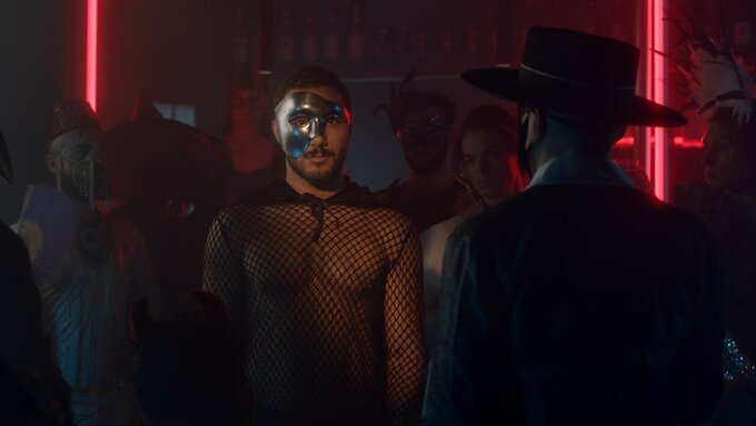 A masked man in a mesh top stands in a crowded nightclub staring into the frame. The background is bathed in neon light.