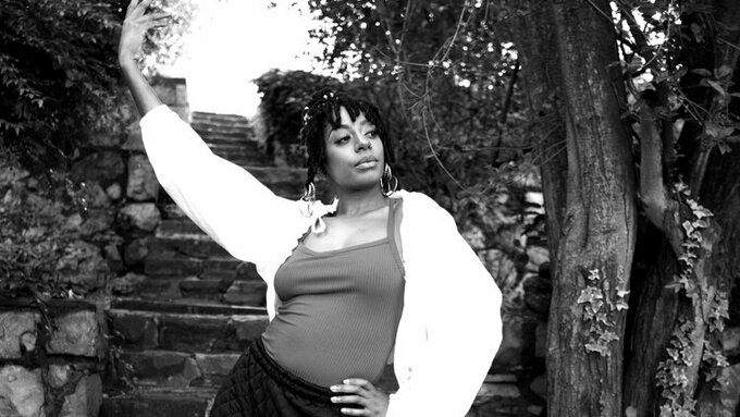 A black femme stands at the foot of a set of stone stairs. One arm is raised, the other holds one side of her hips.