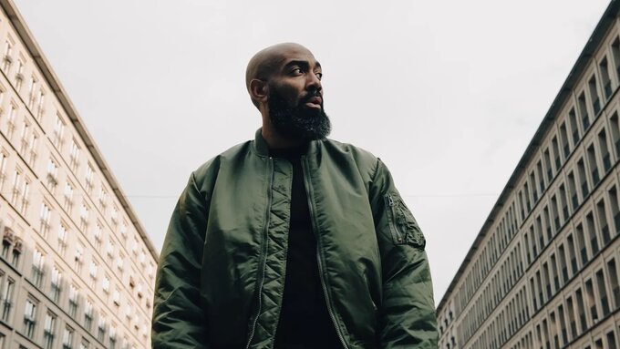 A photograph of Murkage Dave, he wears a green coat and stands between rows of buildings.