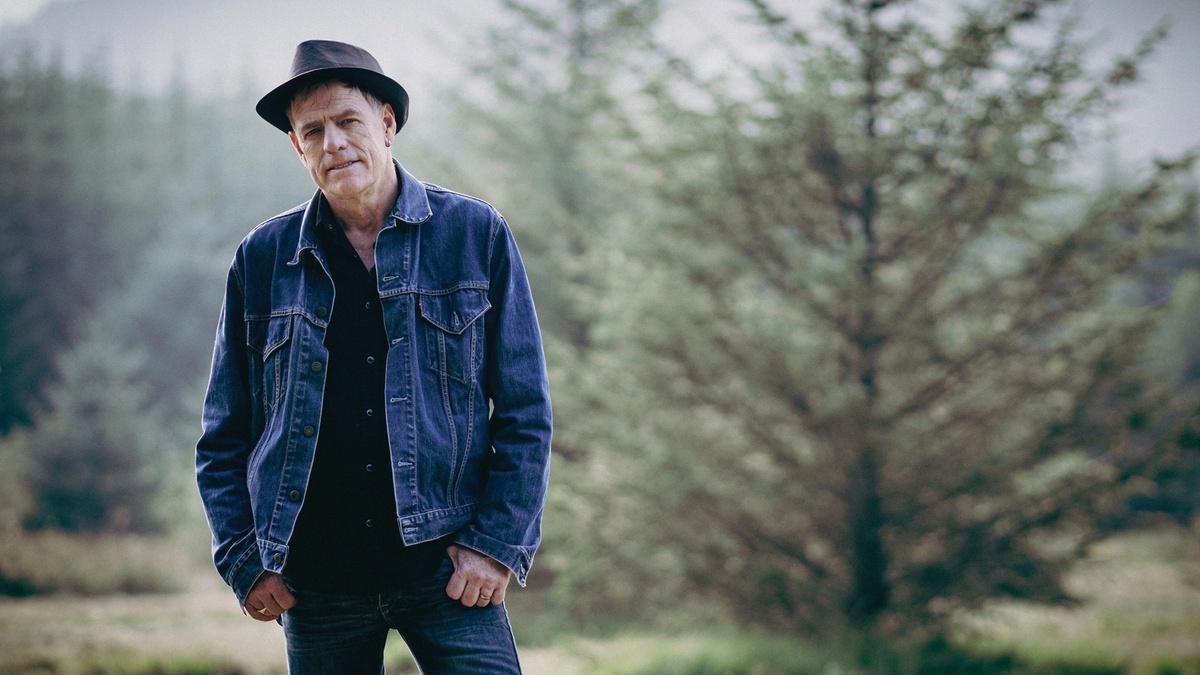 Martyn Joseph stands wearing denim against a green forest.