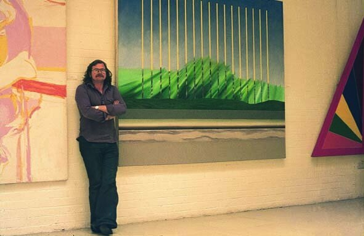 A man with long hair and a moustache stands besides a painting evocative of a landscape in green, blue, and grey.