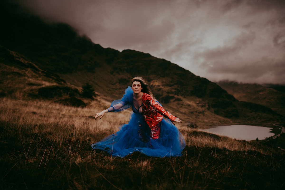 A person in a red and blue dress performing outside amongst green hills and a lake.
