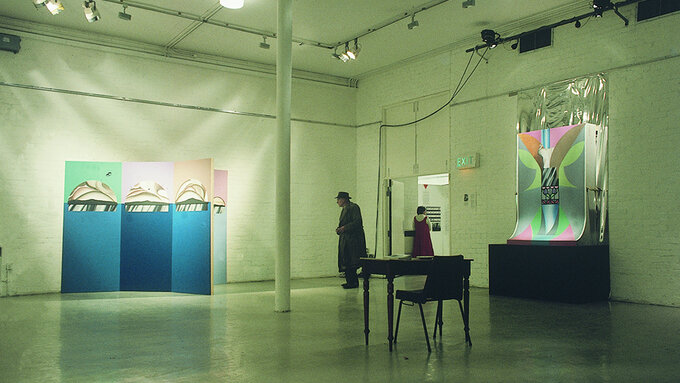 A view of a gallery with two large geometric paintings on each wall and an older man looking at one of them.