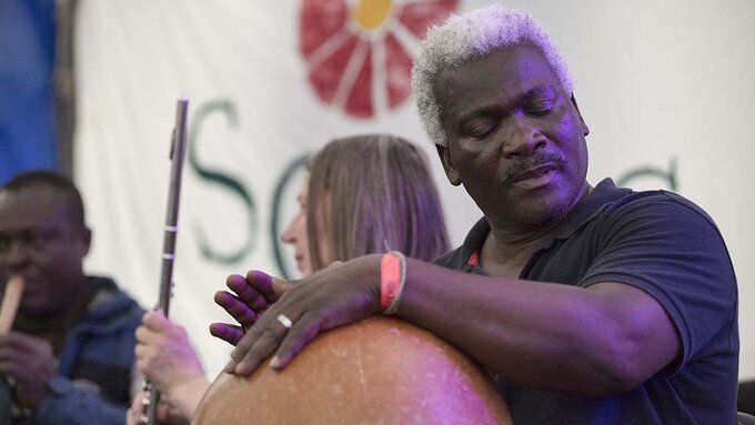 Gameli Tordzro taps a traditional percussion instrument mid-performance, with other musicians in the background.