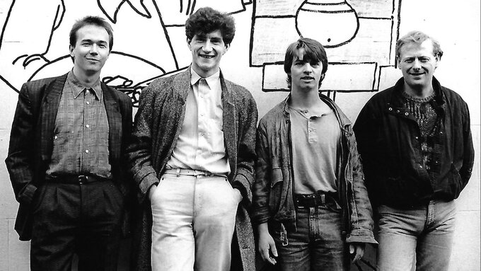 A black and white photo of 4 men smiling and standing in front of a mural of a painter painting a flower pot.