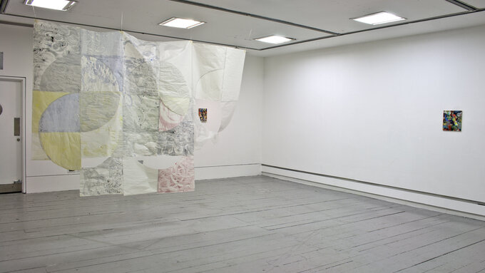 Install view of the show, a geometric quilt hangs from the ceiling and a small painting sits on the far right wall.