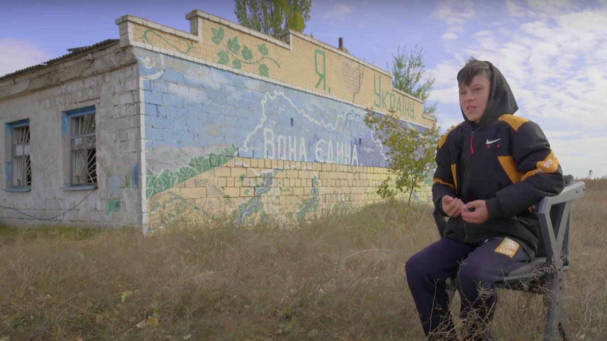 A teen boy sat in a chair in an empty field. In front of a house with graffiti (it reads 'I love Ukraine' in Ukrainian).