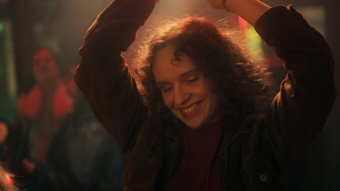 A woman with curly hair, dancing and smiling, against a blurred background of a warmly lit space with several people.