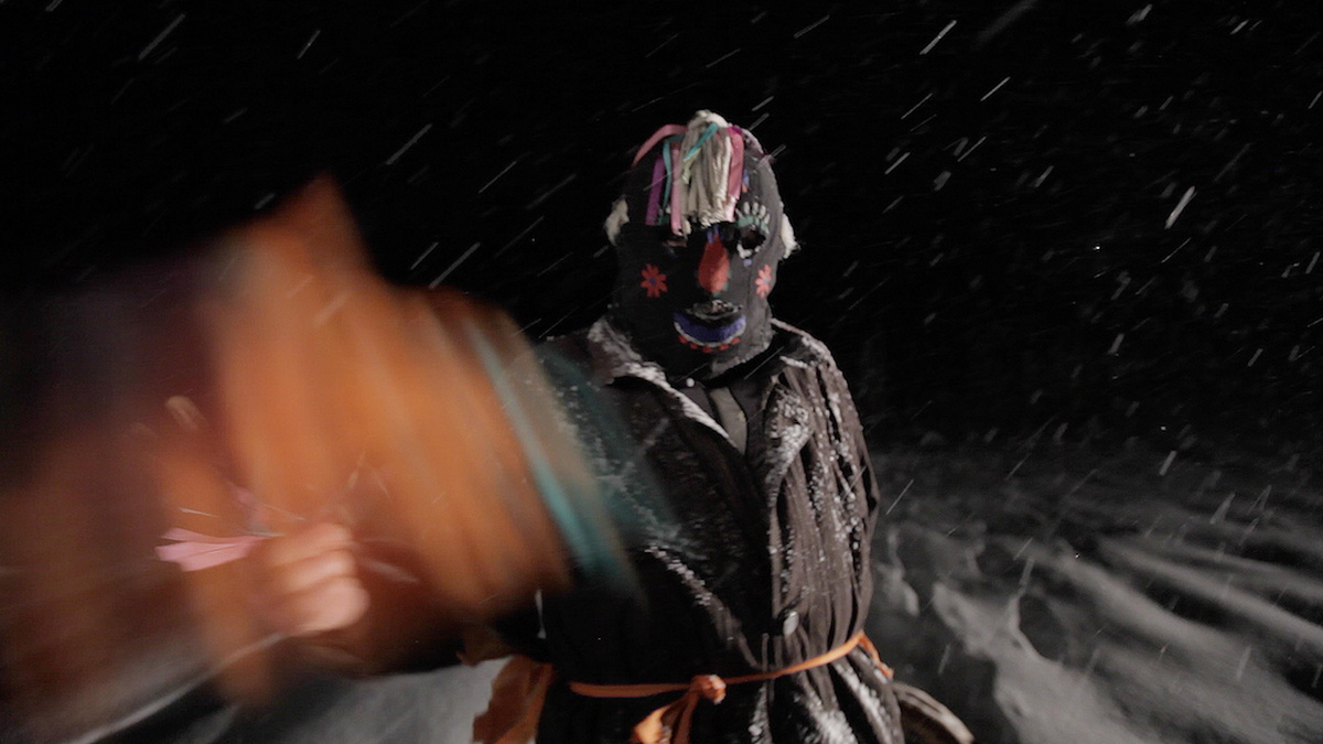 A man wearing a painted folk mask is throwing something bright in the direction of the camera, during a snowy night.