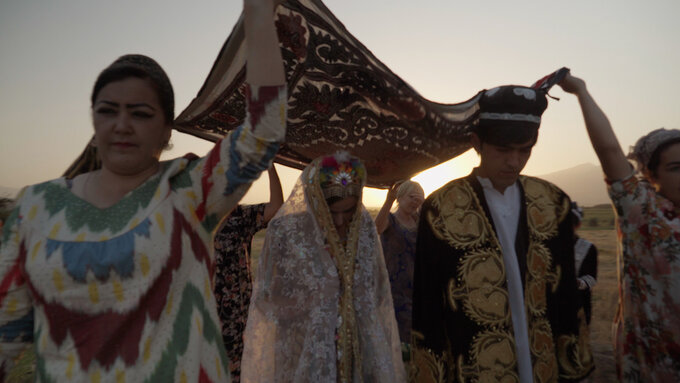 Six people, wearing colourful traditional Tajik garments hold a decorated fabric over a woman's head.