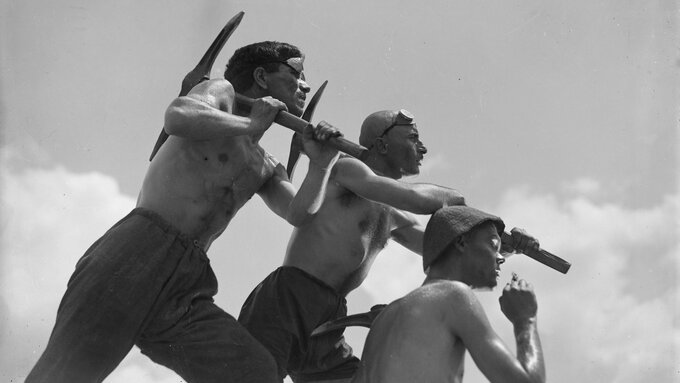 A black-and-white still of three topless men with pickaxes, each with a determined but curious look.