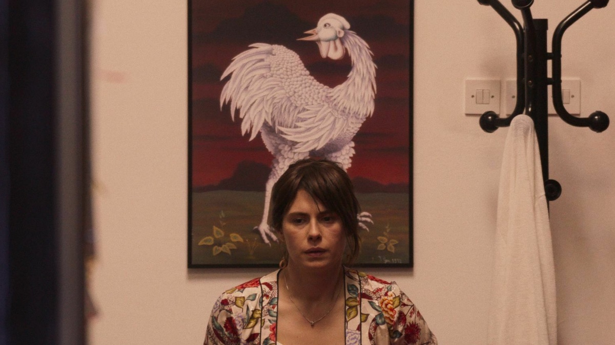 A woman with an exhausted facial expression is sitting in front of a folk painting of a white rooster.