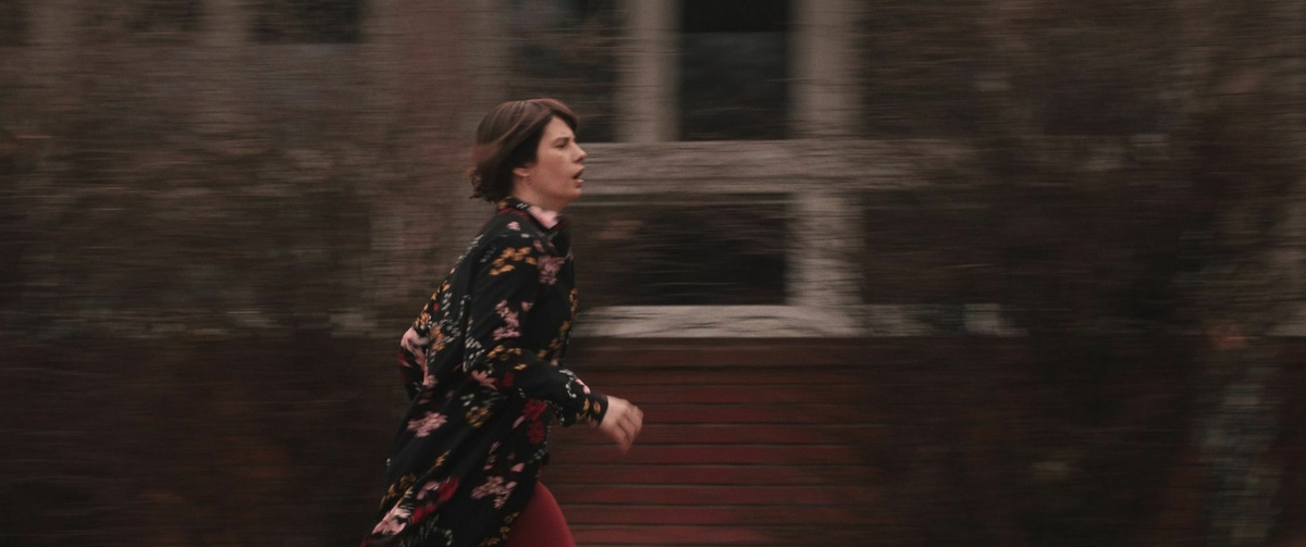 A visibly stressed woman is running towards the right edge of the shot, wearing a black floral cardigan and red trousers