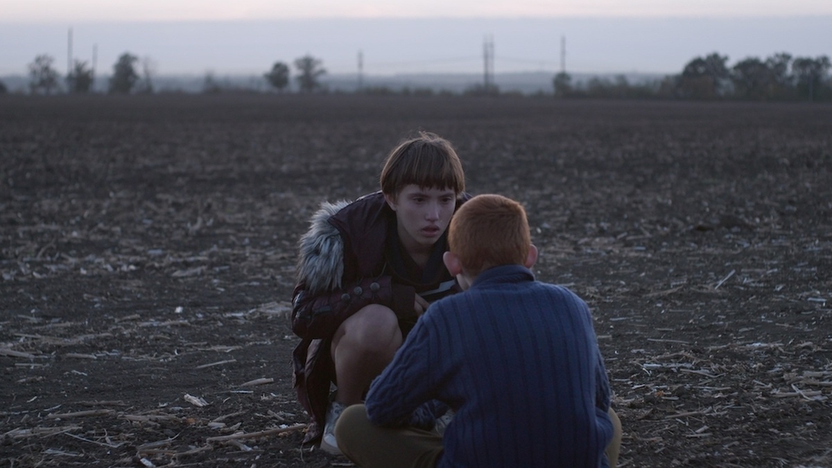 Two children are squatting on the ground, looking at each other, in the middle of farmland, as the sky is getting dark.