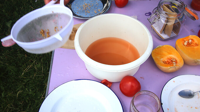 A lilac tabletop holding some bowls, plates, tomatoes, a halved squash and a sieve of seeds.