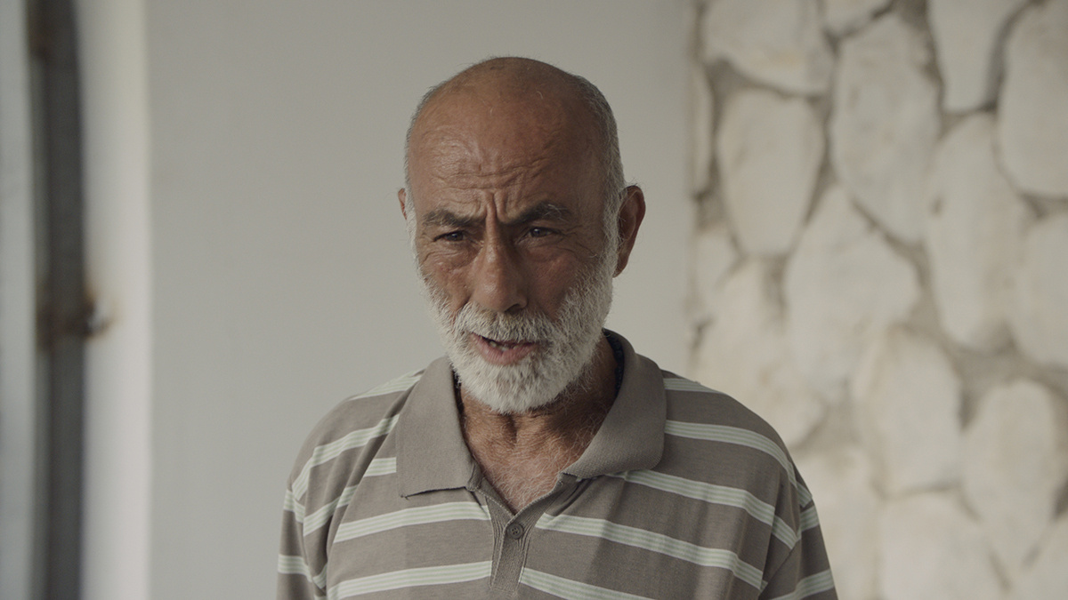 An elderly man with a white beard and striped shirt speaks to the camera. He is in a building with white and stone walls