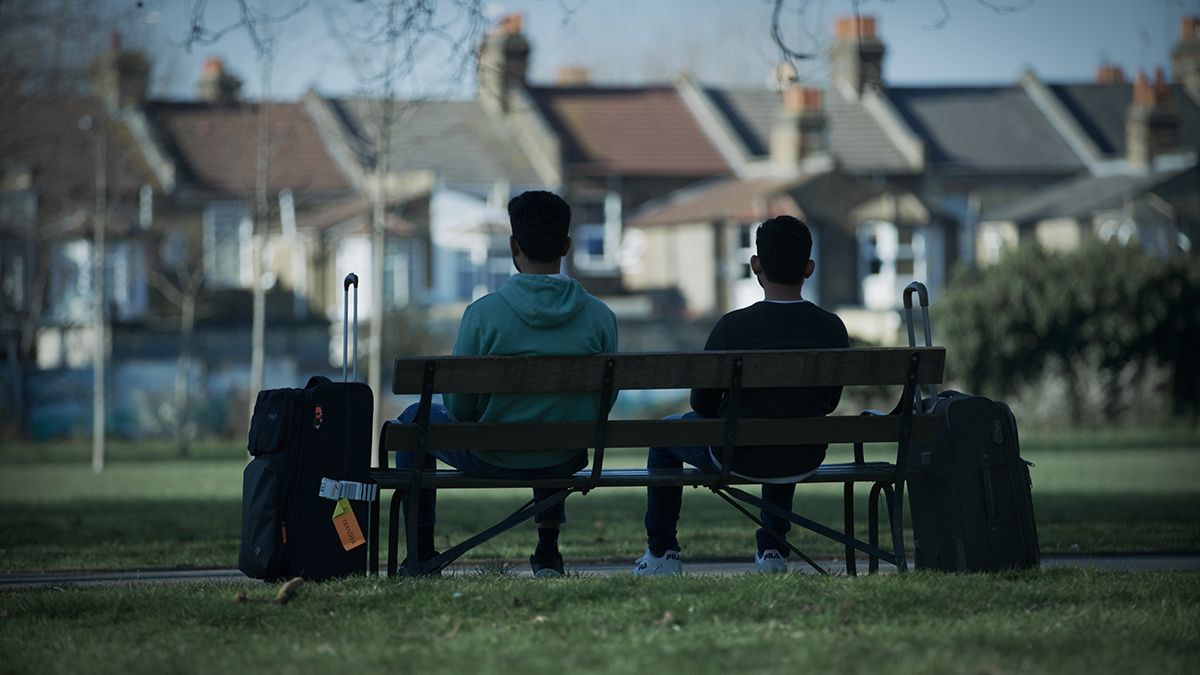 Two people on a park-bench their backs to the camera, with houses in the background and suitcases by their sides.