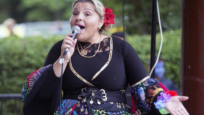 A photo of Sonia in traditional Romani dress, speaking into a microphone and gesturing openly towards  the viewer