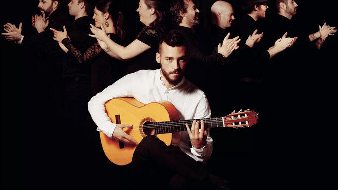 Daniel Martines in the foreground sat holding a guitar, in the background 8 members of the group stand in profile.