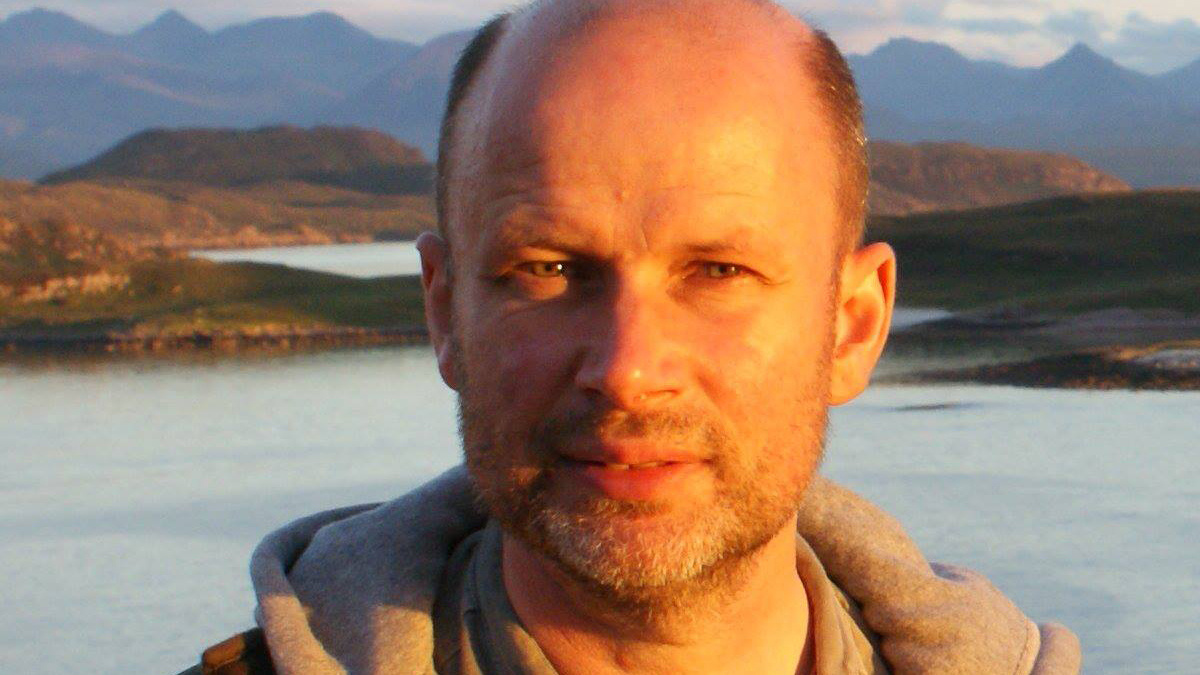 Image of Douglas Thompson with the sun lighting his face. He's outside with a loch and mountains in the background.