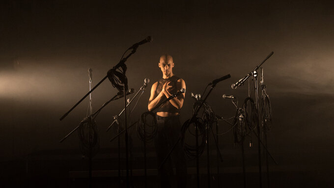 Tiran standing in a dark room; lit by 2 lights from each side of the room. They look at the camera clasping their chest.